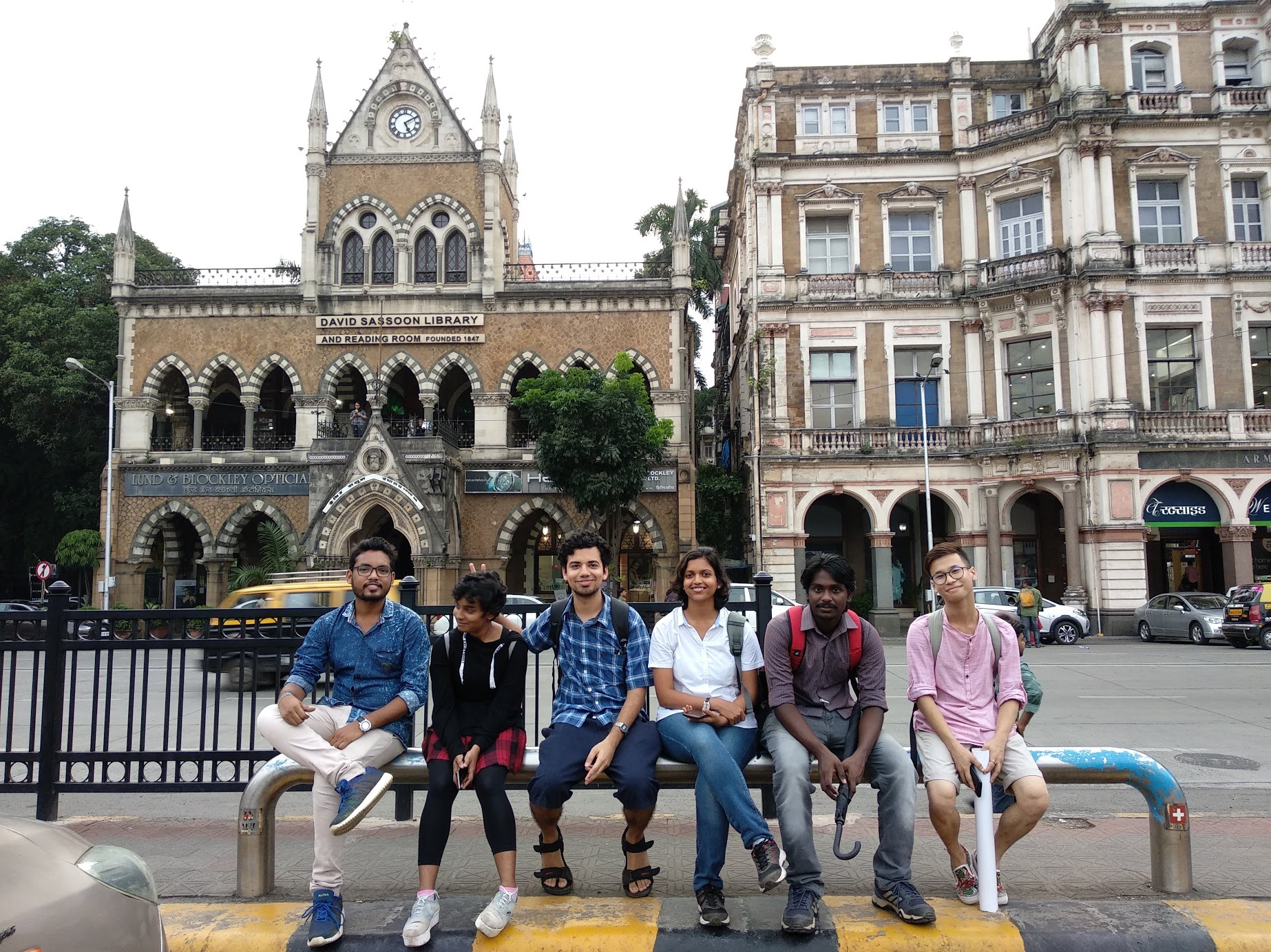 6 people sitting on a metallic tube. Behind them is a busy road and the David Sassoon Library building.